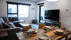 Ein modernes Wohnzimmer mit einer Couch, einem Esstisch gedeckt mit Tellern und Gläsern, einem Fernseher und großen Fenstern.