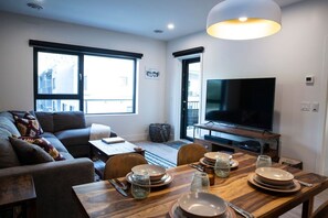 A modern living room with a sofa, a dining table set with plates and glasses, a television, and large windows. - Revy Rendezvous | Private Hot Tub & Mountain Views (Revelstoke)