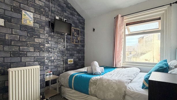 A bedroom with a brick wall, a radiator, a bed with blue and white bedding, a window with curtains, and a small shelf with items. - Suncrest Hotel (Blackpool)