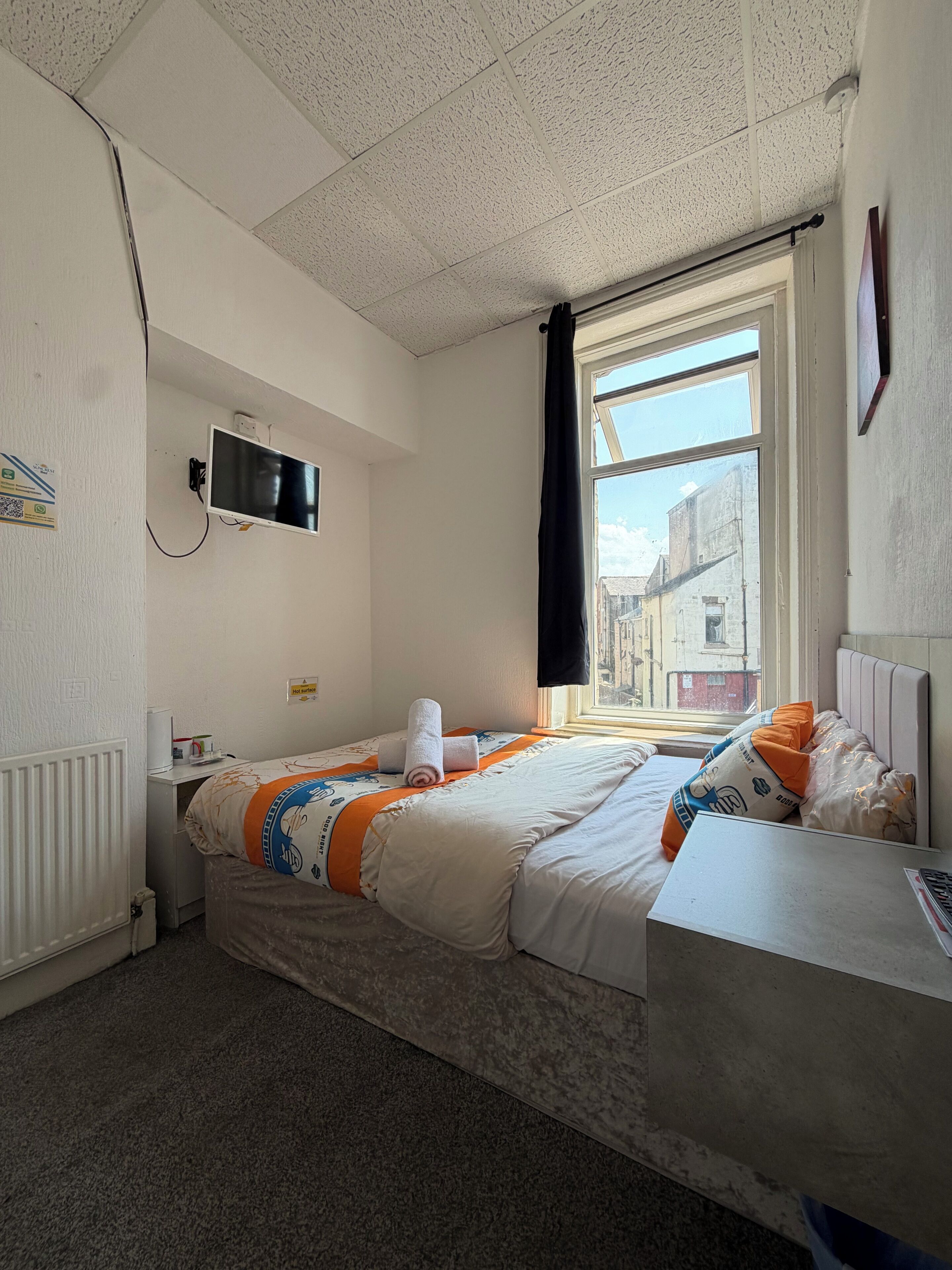 A hotel room with a bed, a desk, a TV, and a window with a view of buildings.