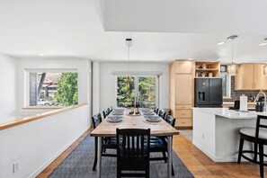 Eine moderne Küche mit einer zentralen Kochinsel, einem Essbereich mit einem langen Tisch und großen Fenstern mit Blick ins Freie.