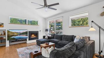 A modern living room with a fireplace, a large flat-screen TV, a gray sectional sofa, and a ceiling fan.