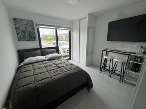Une chambre à coucher comprenant un lit, un tableau, un téléviseur, un bureau avec des tabourets et une fenêtre donnant sur des immeubles.