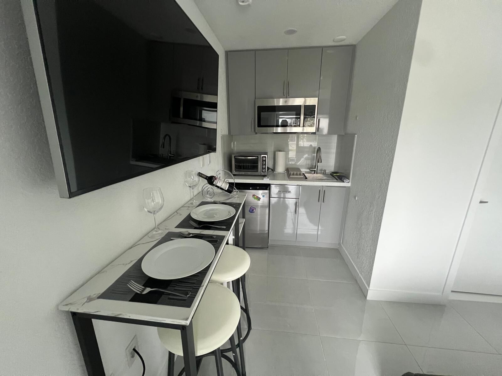 A compact kitchen with a dining area, including a table with chairs, a microwave, a refrigerator, and a large mirror.