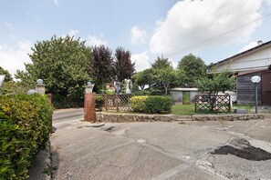 A gated driveway leading to a residential area with a stone wall, a metal gate, and a house with a balcony.