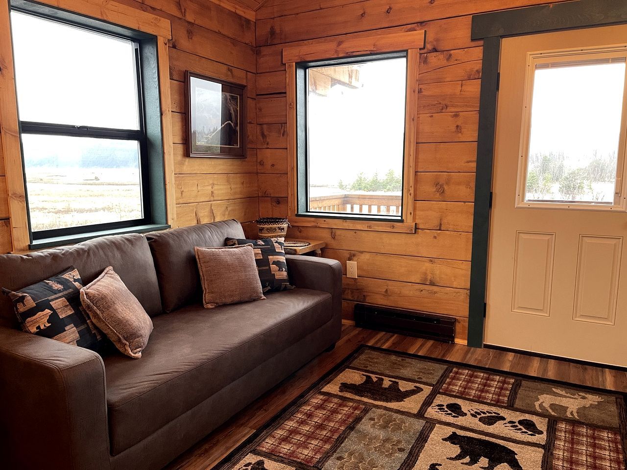 A wooden cabin interior with a sofa, framed pictures, and a rug with animal prints.