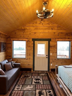 Une chambre située dans une cabane en rondins, avec un plafond en bois, un lustre, un lit, un canapé et deux fenêtres.