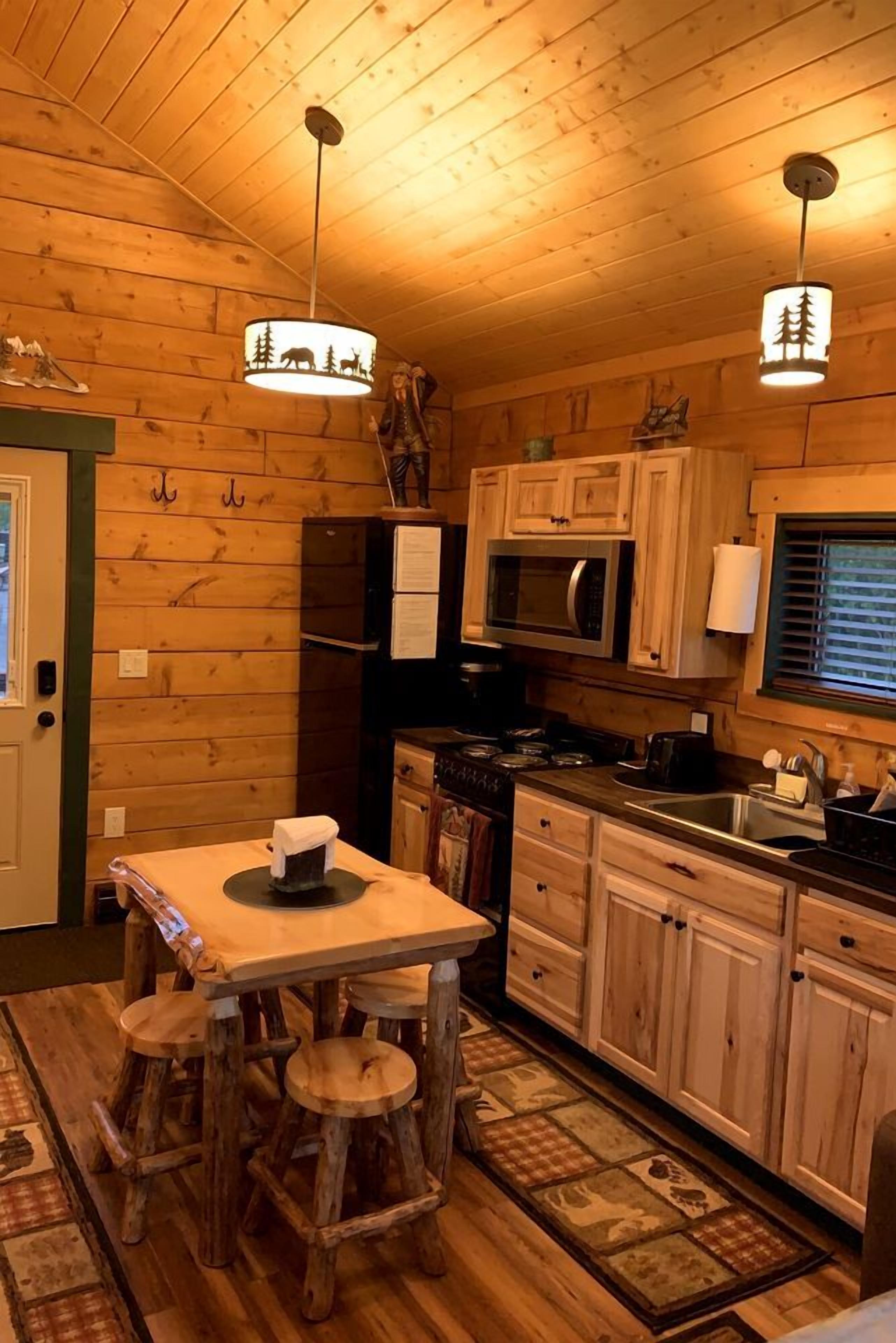 L'intérieur d'un chalet en bois comprend un coin cuisine équipé d'une table en bois, de tabourets, d'un réfrigérateur, d'un four à micro-ondes et d'une cuisinière.