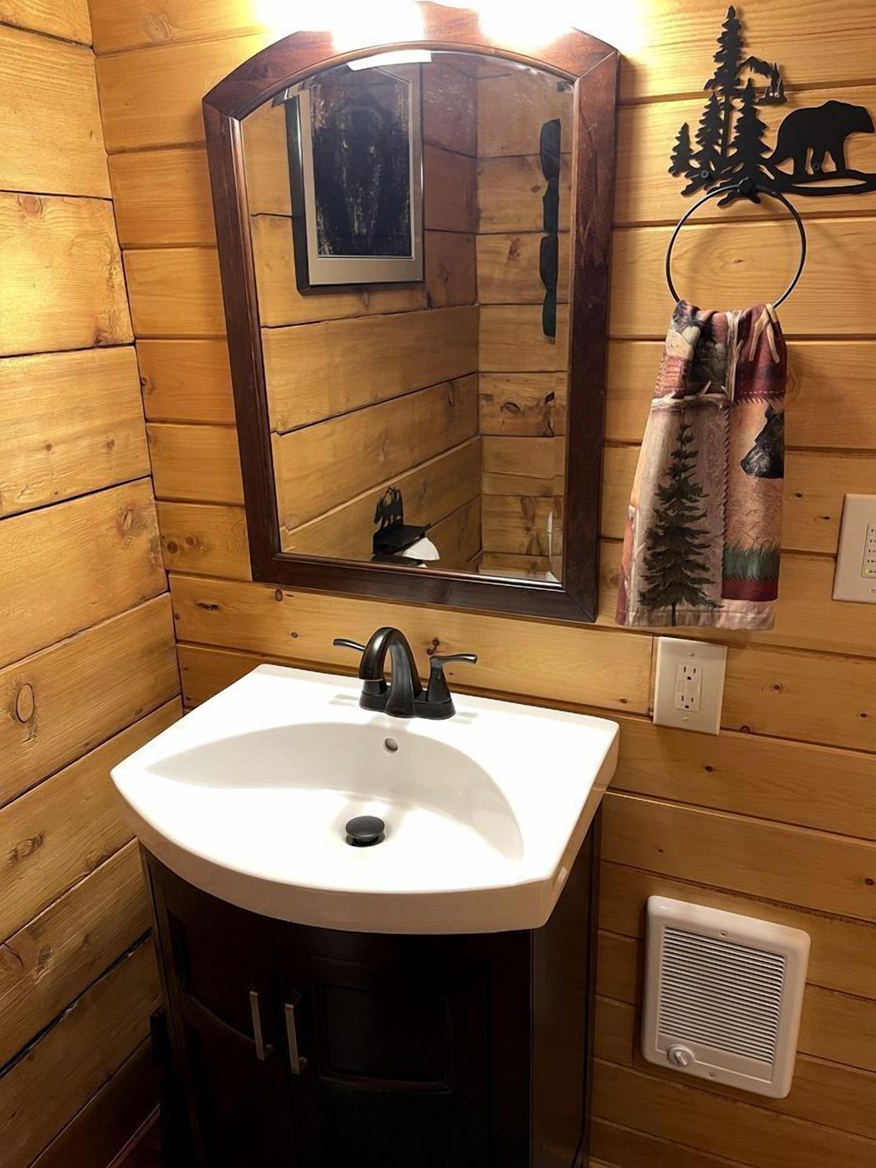 A wooden cabin-style bathroom with a white sink, a mirror, a towel rack with a forest-themed towel, and a vent.