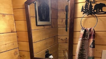 A wooden cabin-style bathroom with a white sink, a mirror, a towel rack with a forest-themed towel, and a vent.