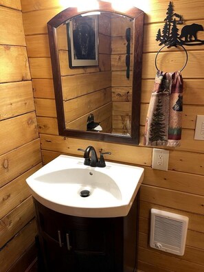 Une salle de bain de style cabane en bois, avec un lavabo blanc, un miroir, un porte-serviettes orné d’une serviette à motifs forestiers et une grille d’aération.