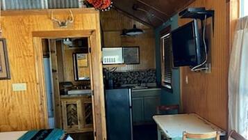 A compact cabin with a bed, a small dining table, a chair, a kitchenette, and a ceiling fan.