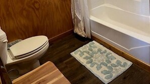 Une salle de bain aux murs lambrissés, équipée de toilettes, d’une douche et d’un petit tapis.