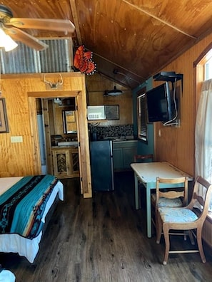 A compact cabin with a bed, a small dining table, a chair, a kitchenette, and a ceiling fan.