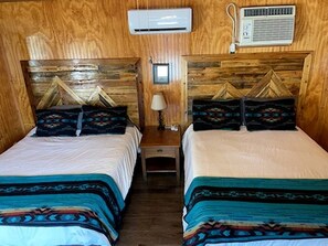 A room with two beds, wooden headboards, an air conditioning unit, a lamp, and a small table.