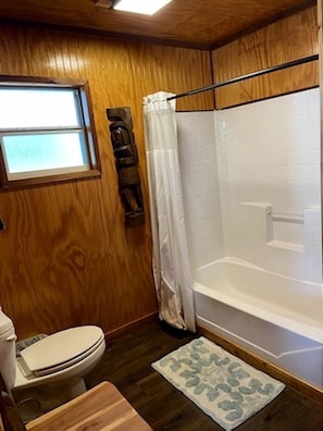 A wooden-paneled bathroom with a toilet, shower, and a small rug.