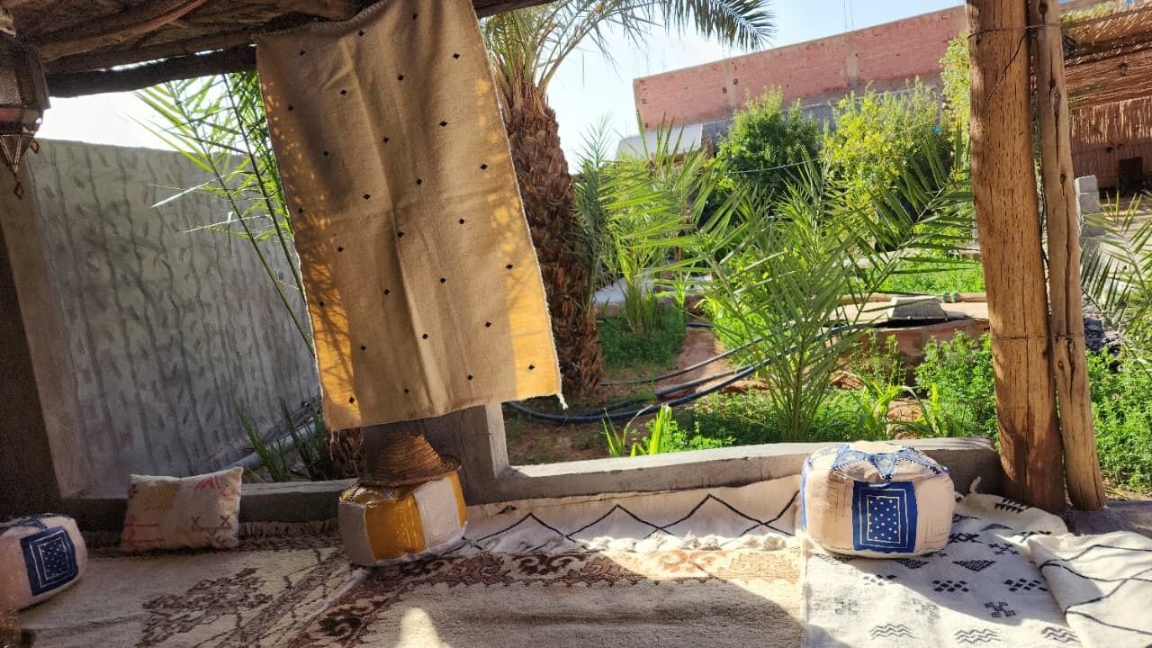 A room with a patterned rug, a hanging curtain, a small table with a candle, and a view of a garden with palm trees.