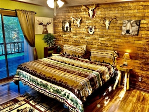 A bedroom with a large bed, a wooden wall with mounted animal skulls, a small side table with a lamp, and a window with curtains.