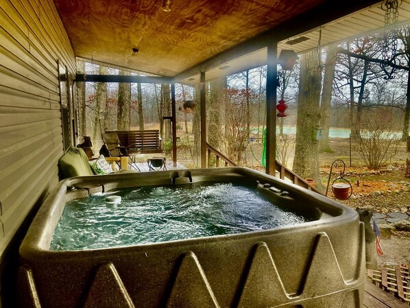 A hot tub in a covered outdoor area with a wooden deck, chairs, and a view of trees and a yard.
