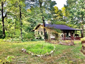 A rustic cabin with a porch, surrounded by a natural landscape with trees and a grassy area.