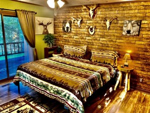 Ein Schlafzimmer mit einem großen Bett, einer Holzwand mit montierten Tierkopfschädeln, einem kleinen Nachttisch mit Lampe und einem Fenster mit Vorhängen.