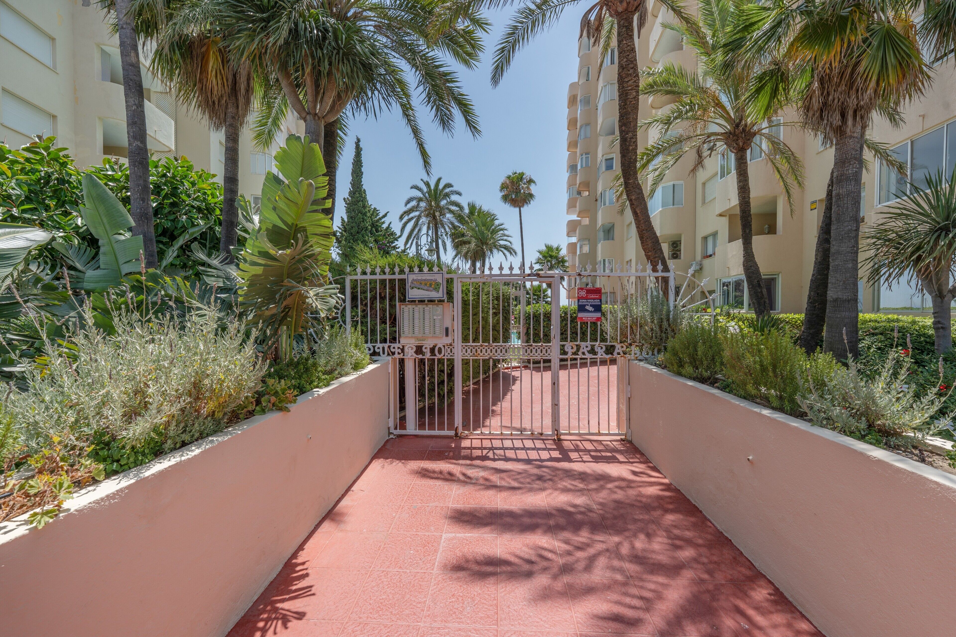 Un cancello d'ingresso recintato con una grata metallica, palme e un edificio con balconi sullo sfondo.