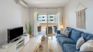 Ein modernes Wohnzimmer mit Flachbildfernseher, einem blauen Sofa, einem Glastisch und Blick auf einen Außenbalkon.
