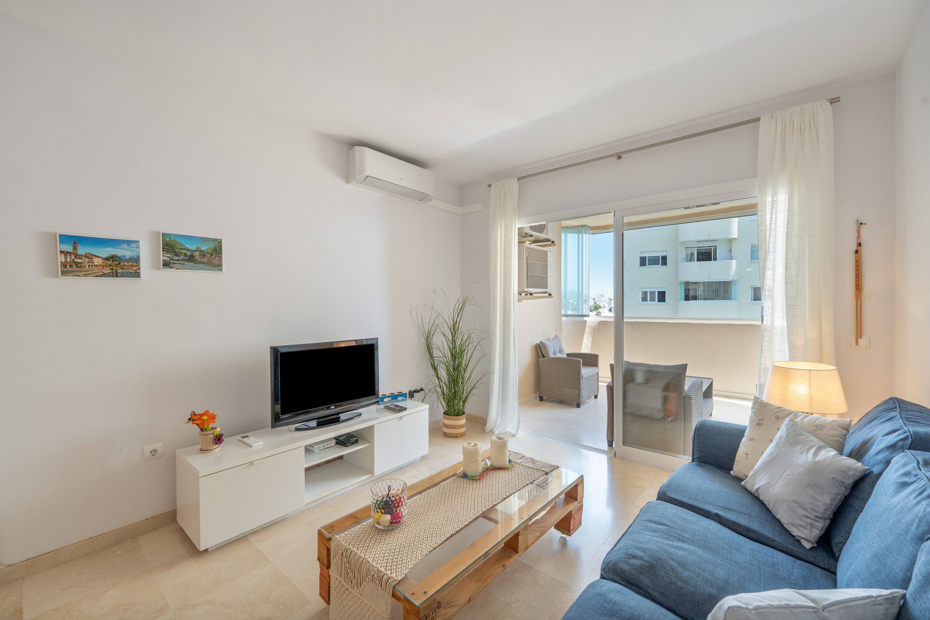 A modern living room with a flat-screen TV, white furniture, a glass coffee table, and a view of a building through a glass door.