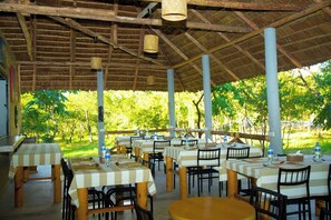Un área de comedor al aire libre con mesas y sillas, techo de paja y vista a la vegetación.