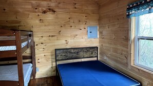 A wooden cabin room with a bunk bed, a single bed, a window, and a small electrical outlet.