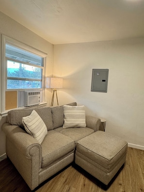 A sectional sofa with pillows, a lamp, a window with blinds, and a thermostat on the wall. - 1-bedroom apartment in Weed (Weed)