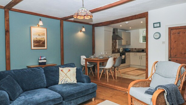 Una sala de estar con un sofá azul, una silla de mimbre, una mesa de comedor con sillas, una cocina al fondo y un techo de madera.