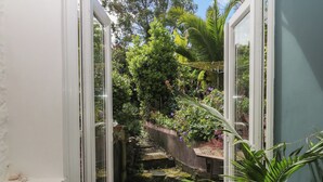 Un estrecho sendero de jardín que conduce a una puerta, rodeado de abundante vegetación y una pequeña fuente de agua.