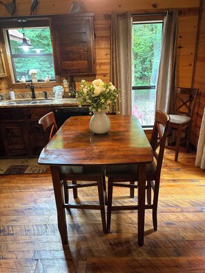 Ein Holzespeisetisch mit Stühlen, eine Vase mit weißen Blumen, eine Küche mit Holzschränken und ein Fenster mit Blick auf Grünflächen.