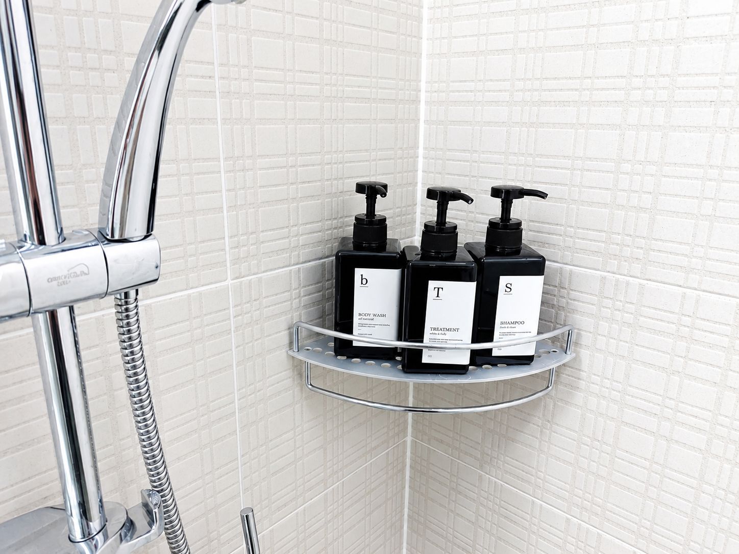 A bathroom with a chrome shower, a white tiled wall, and a shelf holding black bottles labeled 'BODY WASH,' 'TREATMENT,' and 'SHAMPOO.'