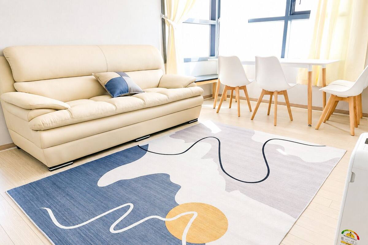Ein modernes Wohnzimmer mit einem beigefarbenen Sofa, einem gemusterten Teppich und einem Essbereich mit weißen Stühlen und einem Tisch.