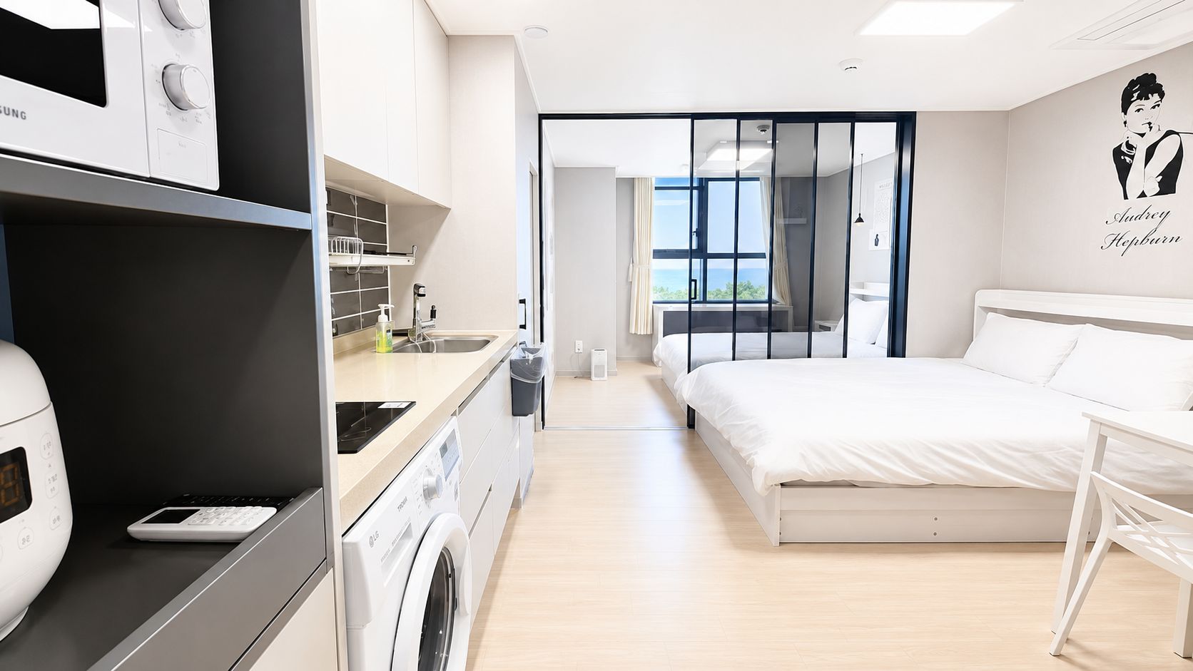 A modern hotel room with a bed, a desk, a washing machine, and a view of the outdoors.
