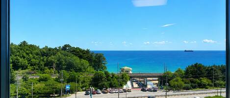 A coastal view from a window, including a road with vehicles, a parking lot, and a distant ship on the water.
