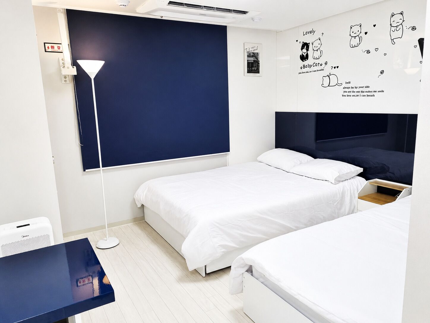 A bedroom with two beds, a desk, a lamp, and a wall with cat illustrations.