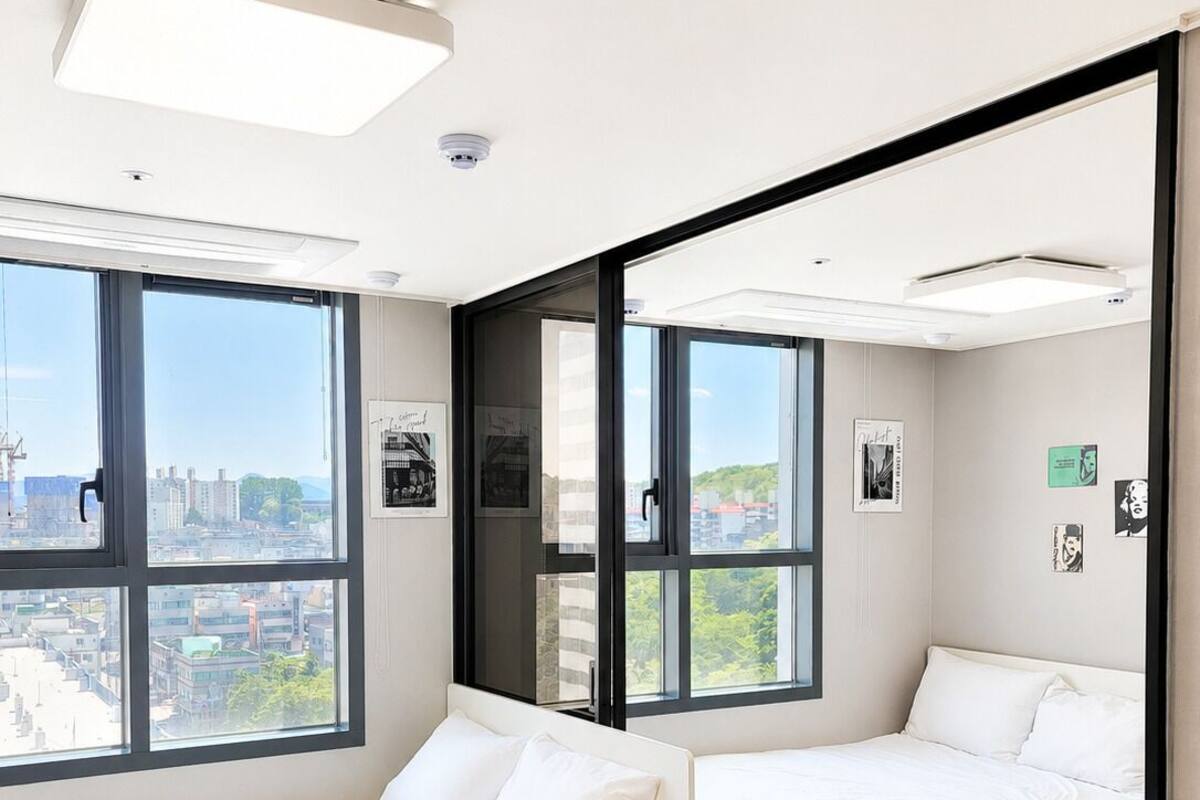 Ein modernes Hotelzimmer mit zwei Betten, großen Fenstern, Blick auf die Stadt und einem Spiegel, der den Raum widerspiegelt.