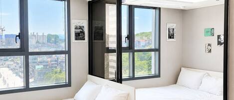 A modern hotel room with two beds, large windows, a city view, and a mirror reflecting the room.