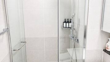 Un baño moderno con ducha de vidrio, lavamanos blanco y un pequeño estante con artículos de aseo.