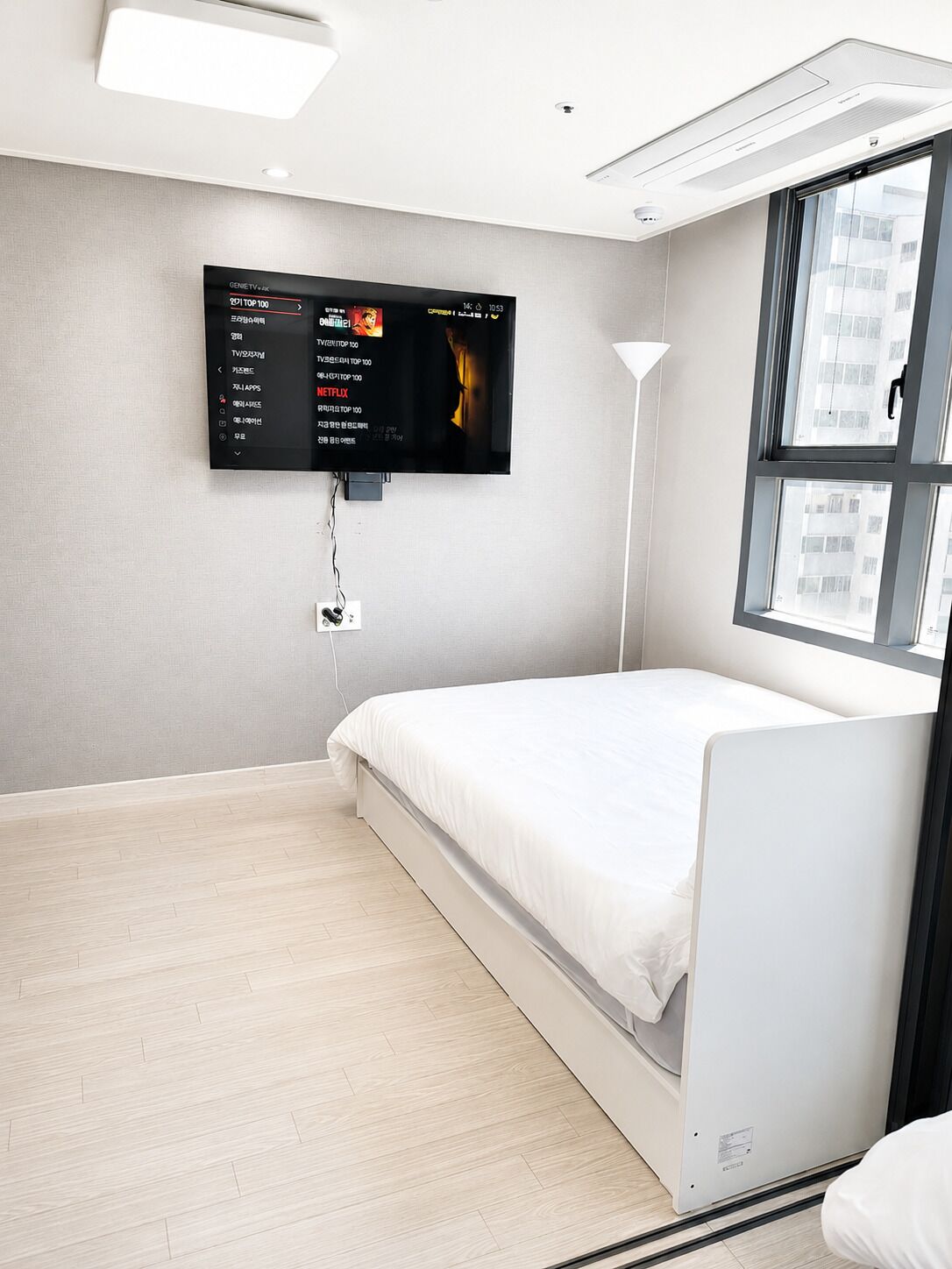 Une chambre d’hôtel moderne, équipée d’un lit, d’un téléviseur fixé au mur qui affiche un programme télévisuel, et d’une fenêtre donnant sur des immeubles.