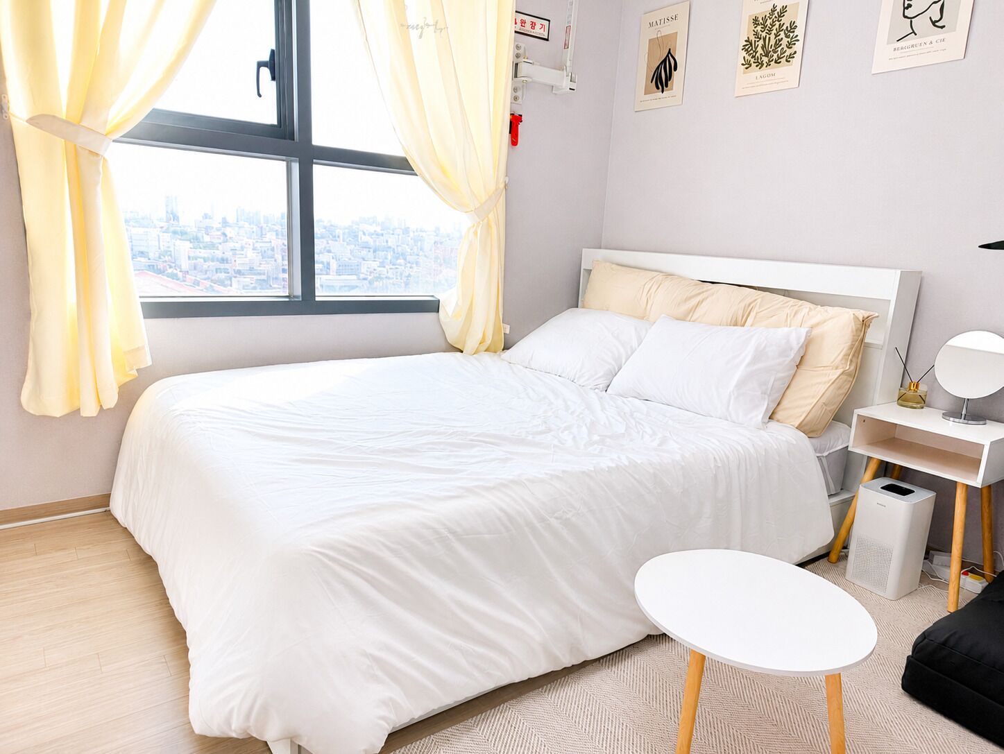 A bedroom with a bed, a bedside table, a lamp, a small round table, and a view of the city through a window.