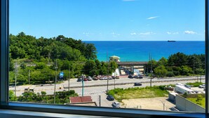 Vista costera desde una ventana, mostrando una carretera con vehículos, un estacionamiento y un barco a lo lejos en el agua.