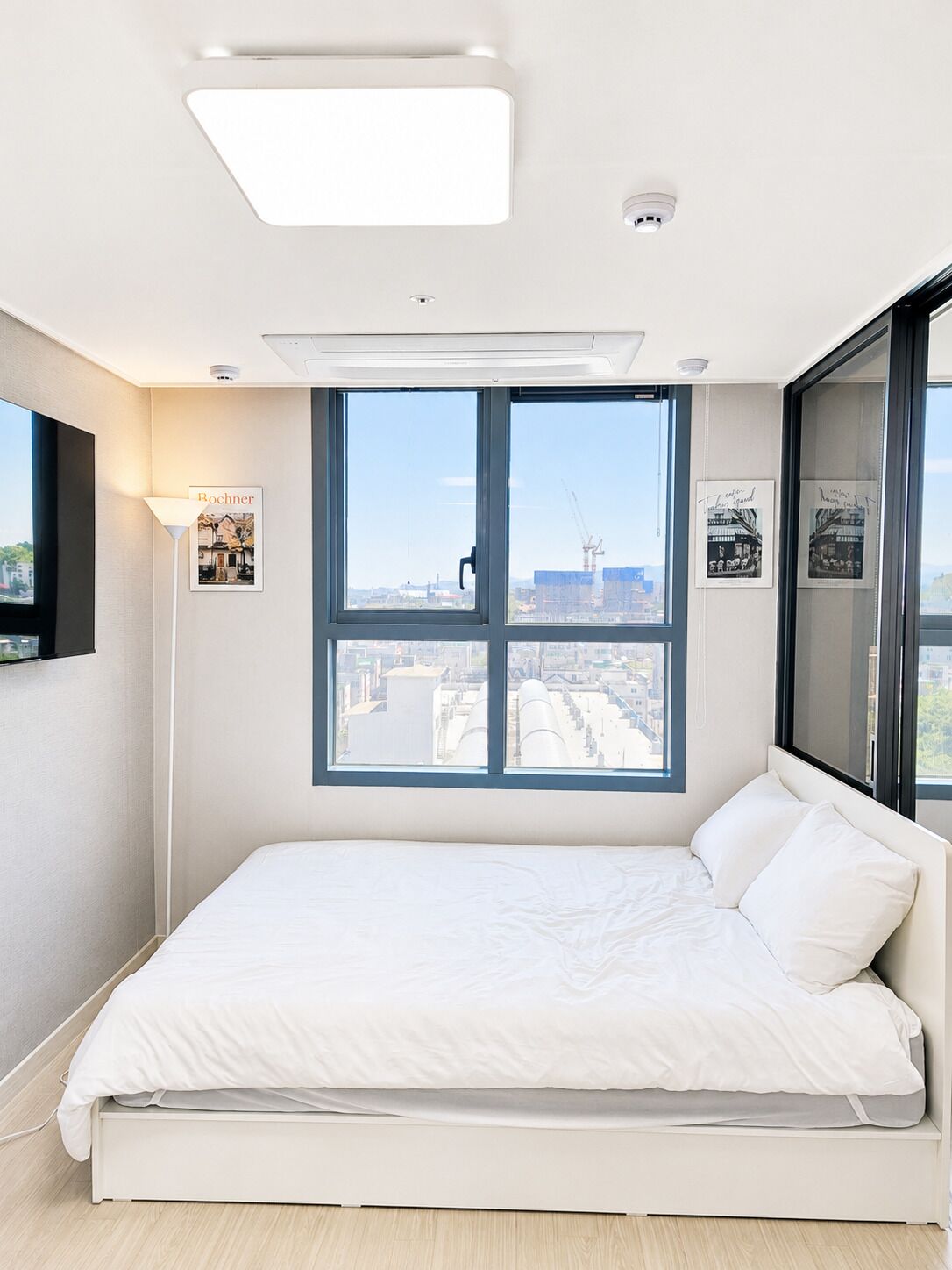A modern bedroom with a large window, a bed, a floor lamp, and a ceiling light.