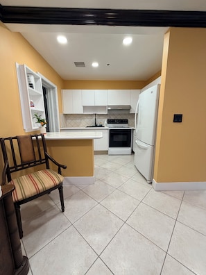 A compact kitchen with white cabinets, a built-in oven, a refrigerator, and a chair with a striped cushion.