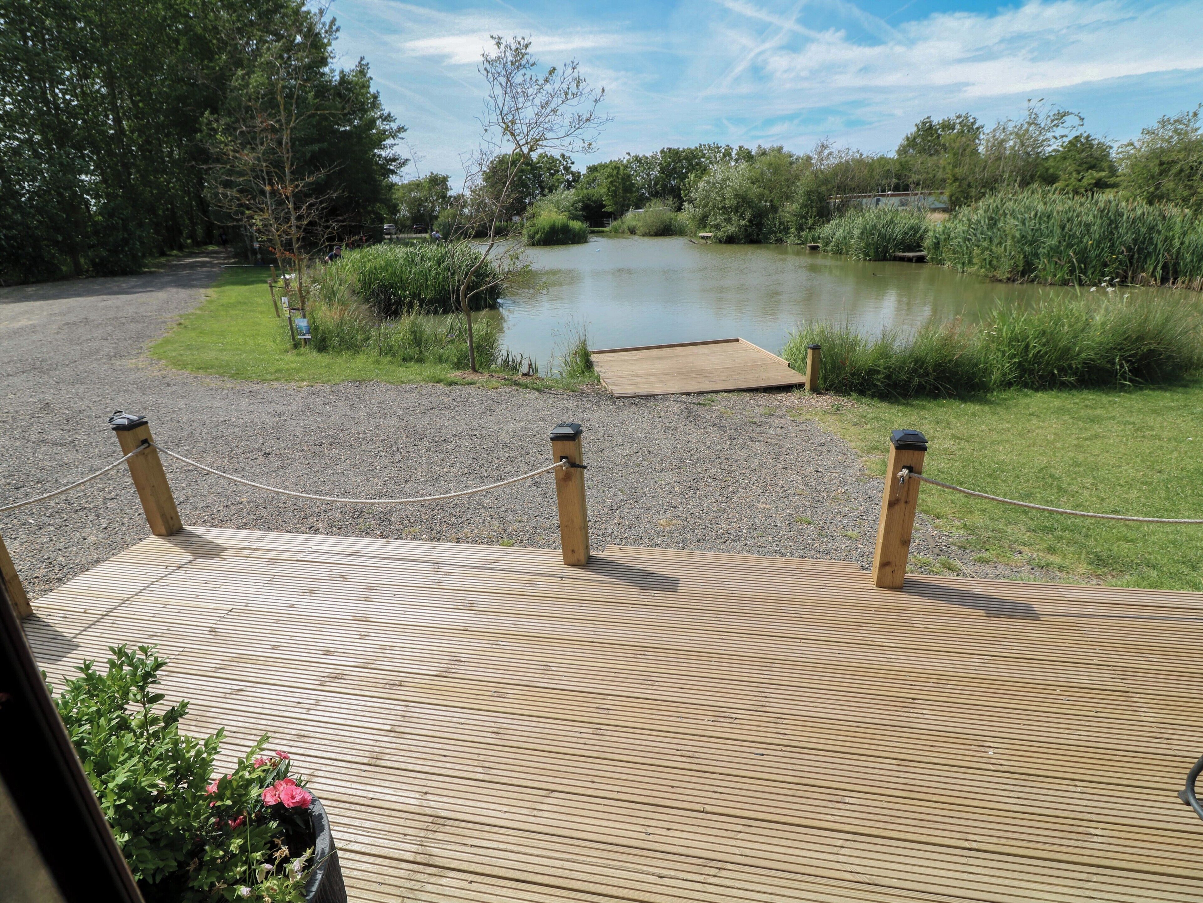 Une terrasse en bois avec une barrière en corde, un chemin de gravier et une étendue d’eau bordée de végétation.