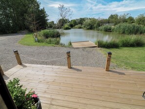 A wooden deck with a rope barrier, a gravel path, and a body of water with vegetation. - Moorhen Lodge (Market Rasen)