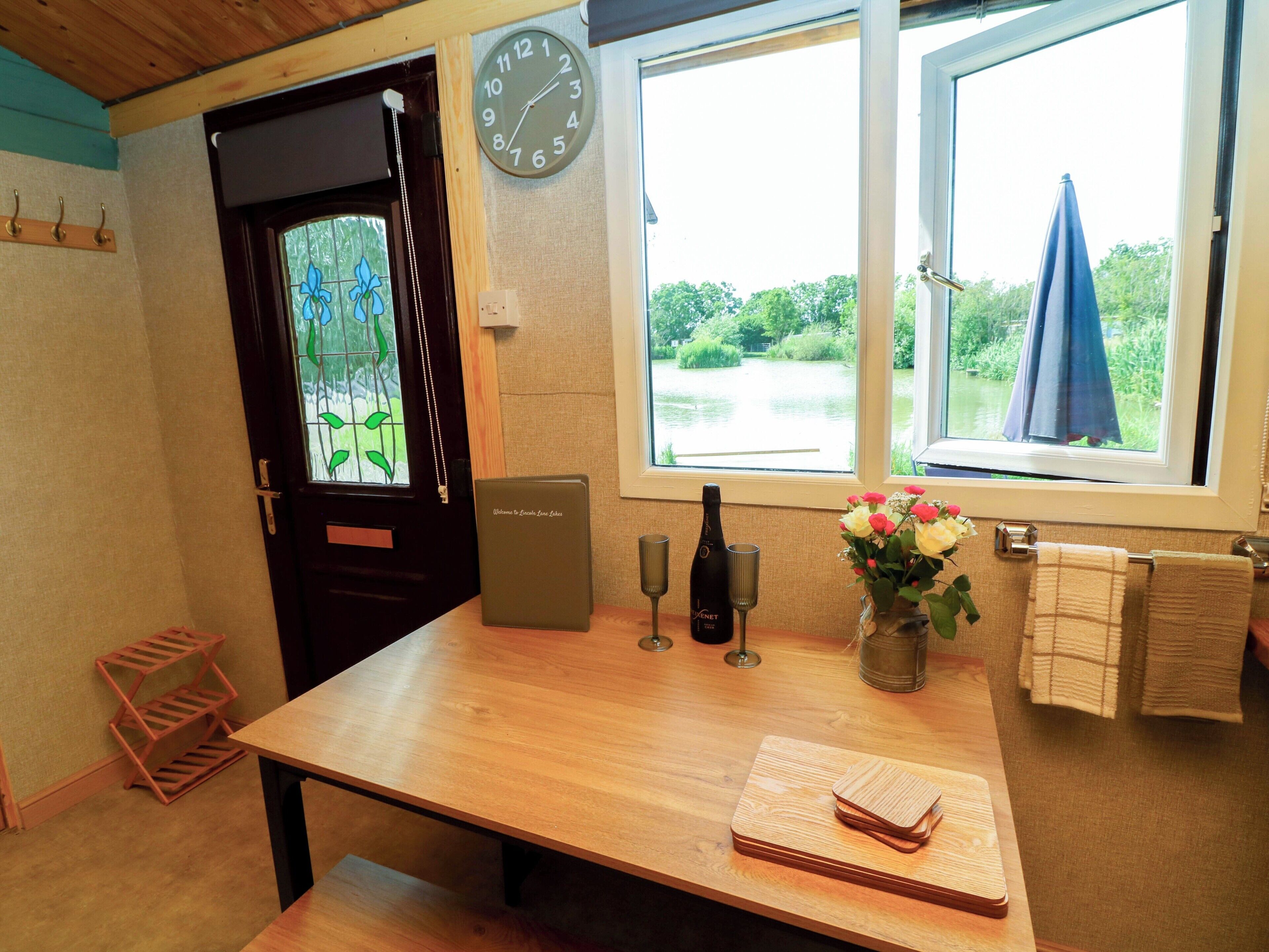A room with a wooden table, a clock, a bottle of champagne, and a window with a view of greenery.